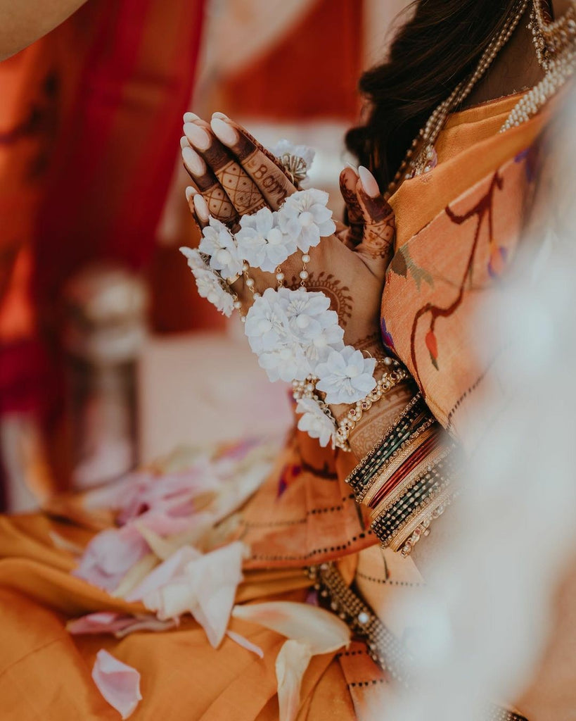 a bride wearing Prune India Signature silk floral haath phool with henna on her hands and sitting with folded hands in a religious ceremony of her wedding 