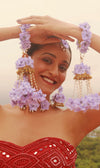 A woman wearing a traditional red dress and floral accessories, including bangles and a headpiece with lavender flowers.