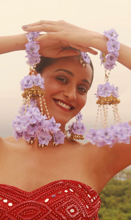 A woman wearing a traditional red dress and floral accessories, including bangles and a headpiece with lavender flowers.
