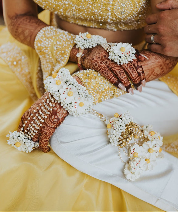 Close-up of hands with henna designs and floral decorations, wearing yellow and white traditional attire.