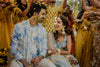 Couple in traditional attire surrounded by confetti and people in colorful clothing.