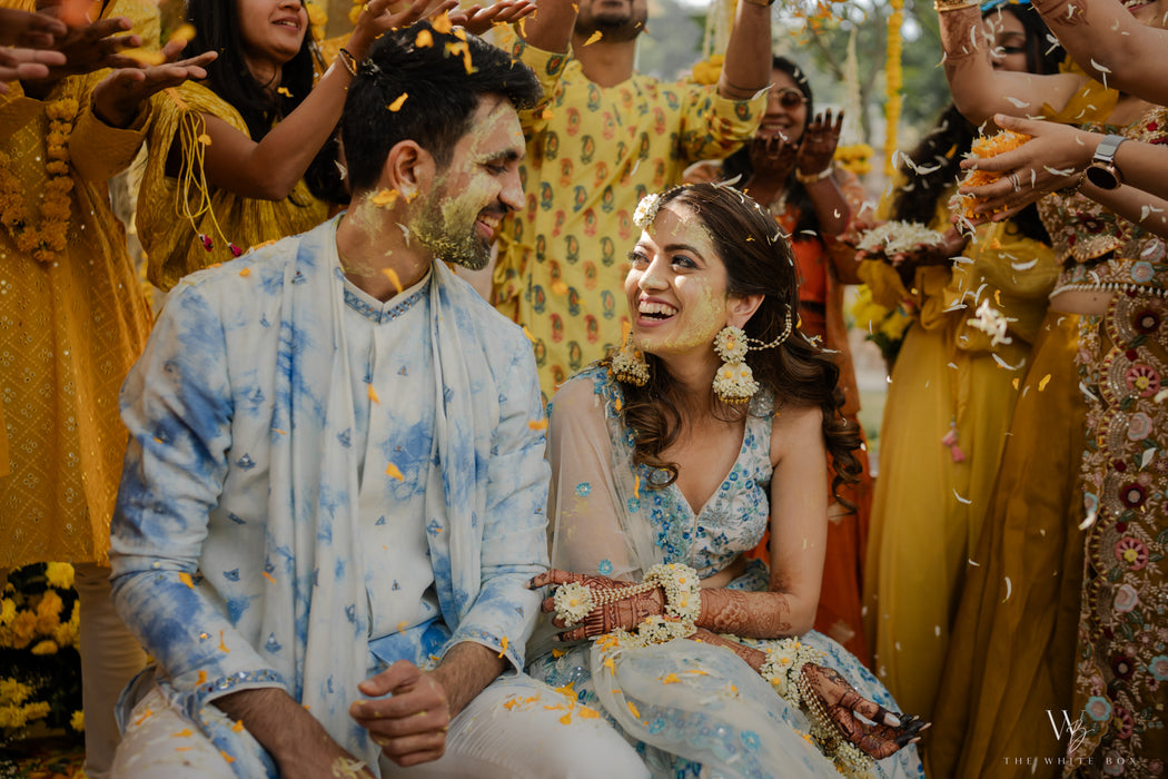 Couple in traditional attire surrounded by confetti and people in colorful clothing.
