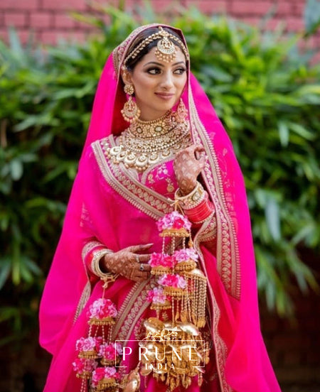 Woman in traditional pink Sabyasachi Bridal Lehnga with jewelry and floral decorations, standing against a green leafy background.