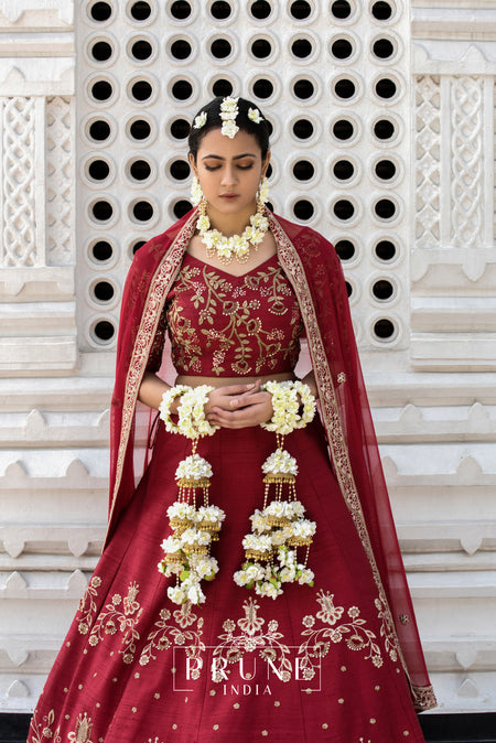 A woman wearing a traditional red bridal lehenga with gold floral embroidery, holding floral accessories, in front of a white geometric patterned backdrop.
